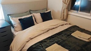 Een netjes opgemaakt bed met een donge deken en meerdere kussens, een nachtkastje met een lamp en een raam met gordijnen.