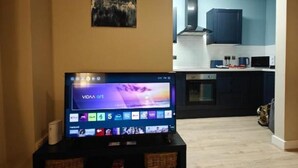 Een moderne woonkamer met een flatscreen televisie op een standaard, een schilderij aan de muur en uitzicht op een keuken met donkere kasten en een magnetron.
