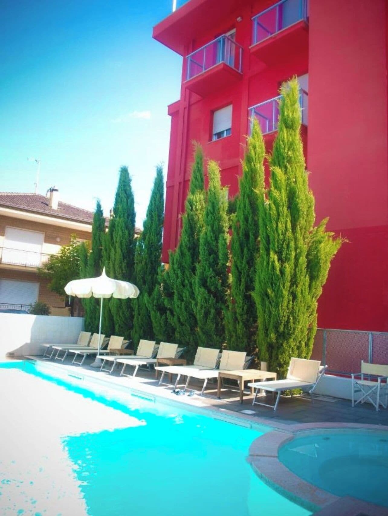 A swimming pool with lounge chairs and an umbrella, surrounded by tall evergreen trees and a red building.