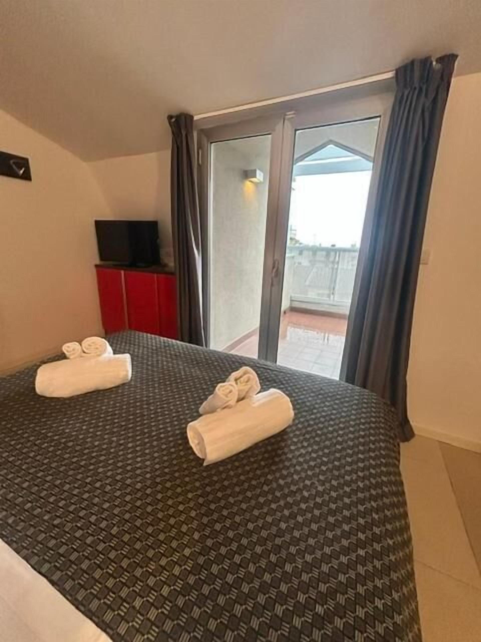 A hotel room with a bed, a TV on a stand, and a balcony with curtains.