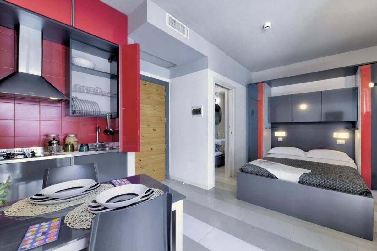Una habitación de hotel moderna con cama, mesitas de noche, una pequeña cocina y una mesa de comedor con platos y cubiertos.