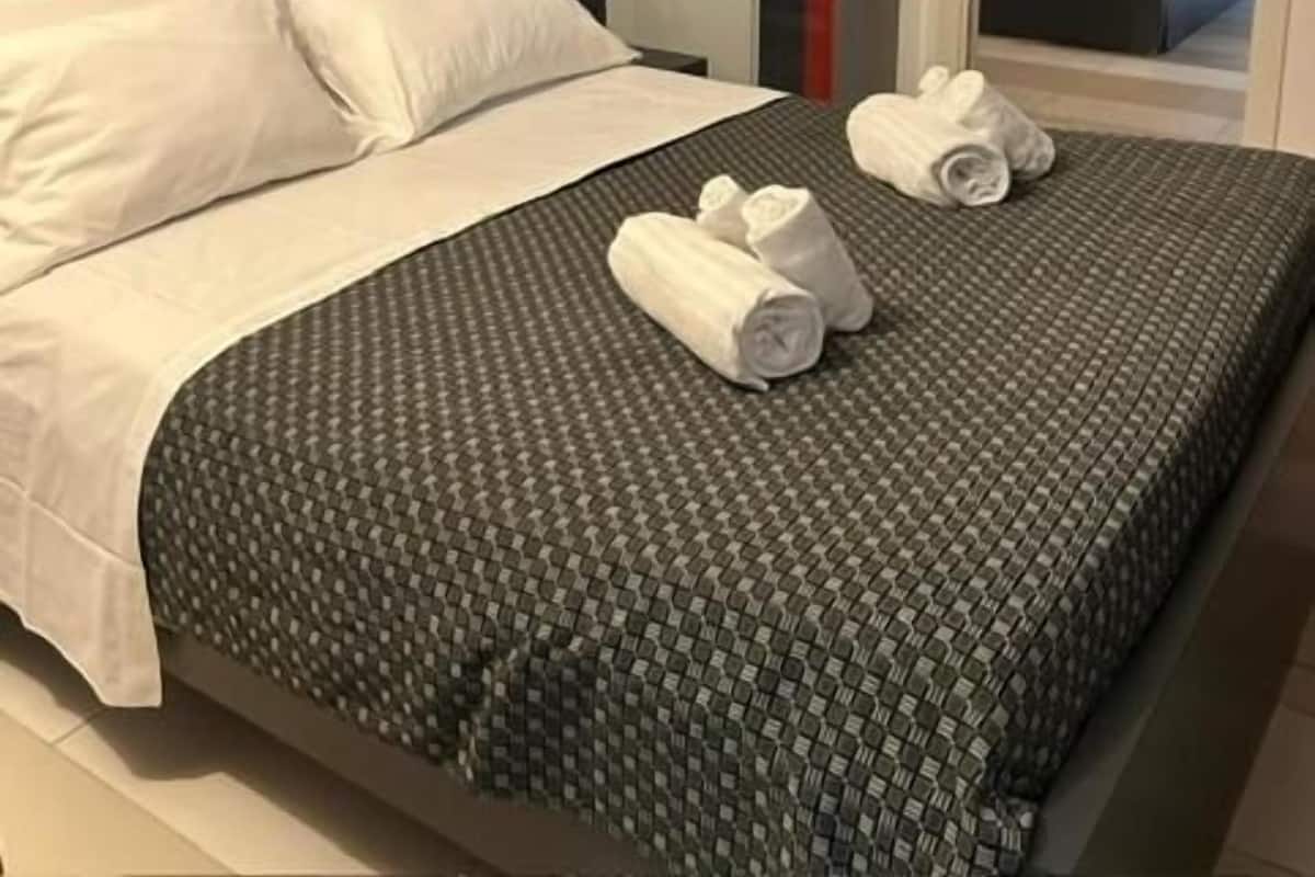 Una habitación de hotel con una cama grande, una colcha de tonos grises con estampado, almohadas blancas y toallas enrolladas sobre la cama.