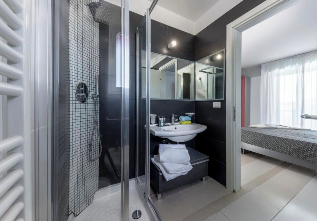 A modern bathroom with a glass shower enclosure, a sink with a mirror, and a bed in the adjacent room.
