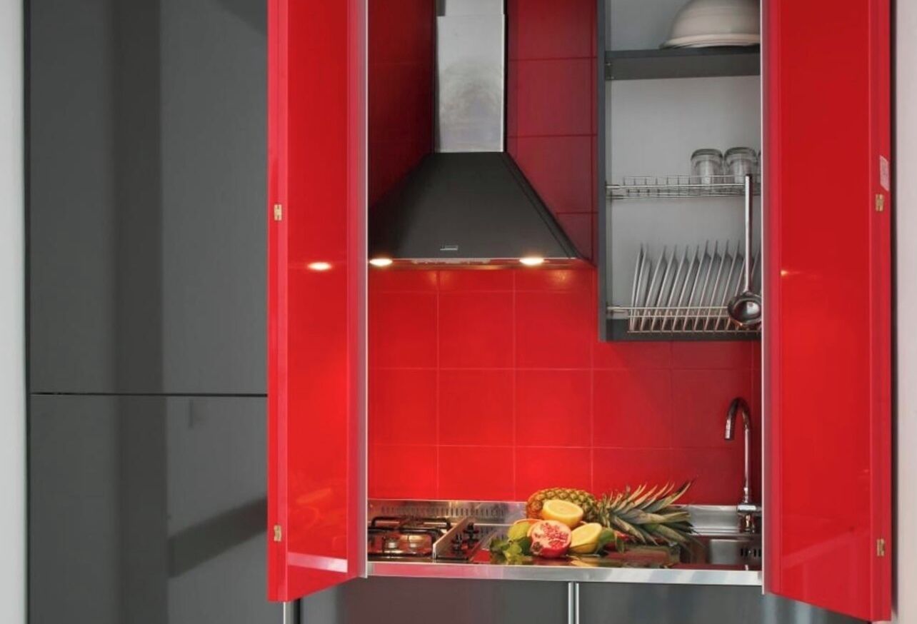 Cozinha compacta com armários vermelhos, pia de aço inoxidável e diversas frutas no balcão.