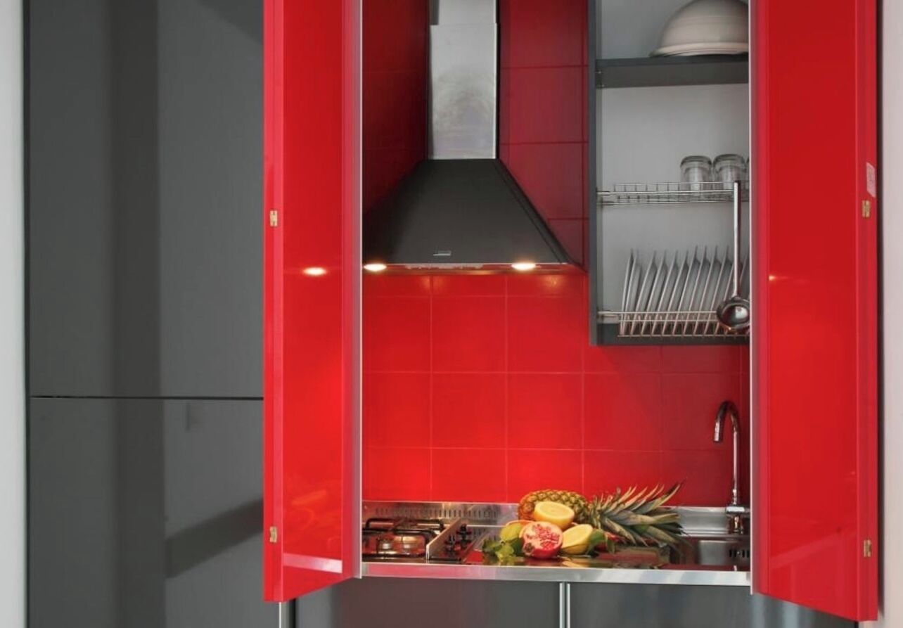 A compact kitchen with red cabinetry, a stainless steel sink, and a variety of fruits on the counter.