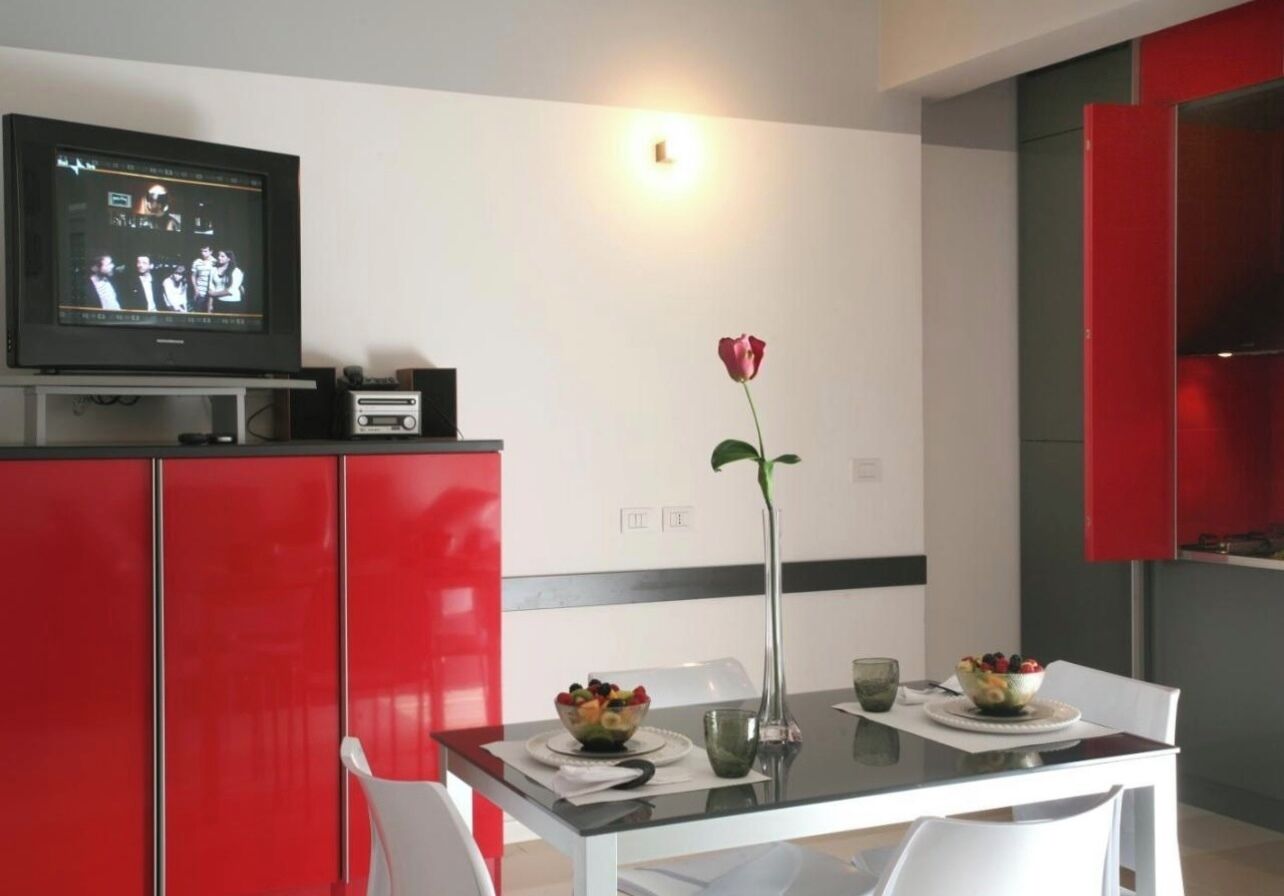 Una cocina moderna con armarios rojos, una mesa de comedor con sillas blancas, un florero con una rosa y un televisor sobre un mueble.