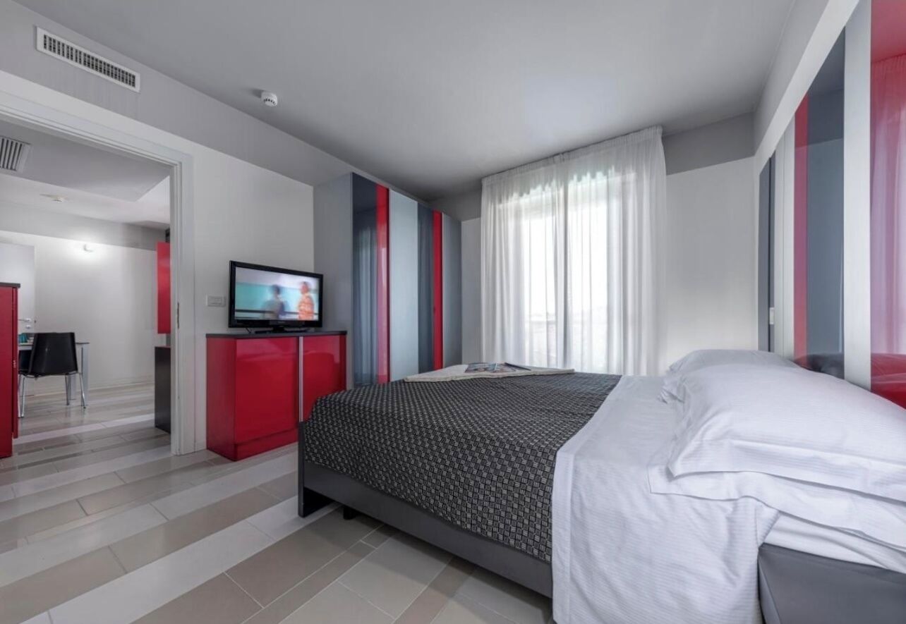 A modern hotel room with a bed, a TV, a desk, and a red wardrobe.