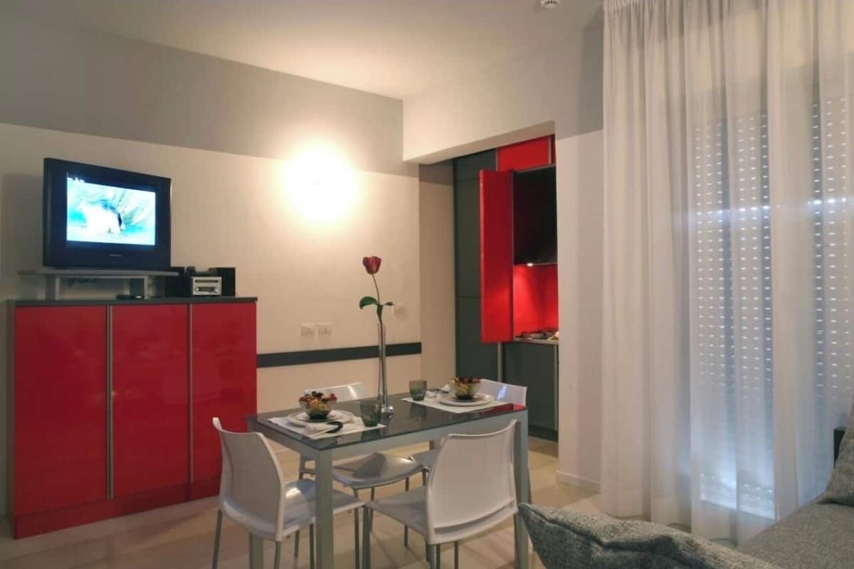 Una sala de estar moderna con un televisor de pantalla plana, una mesa de comedor de vidrio, sillas blancas, un mueble rojo y un amplio ventanal con cortinas translúcidas.