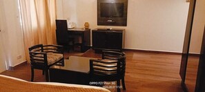 A living room with a wooden floor, a glass coffee table, two upholstered chairs, a television on the wall, and a small table with a lamp. - Hotel Glitz (Jaipur)