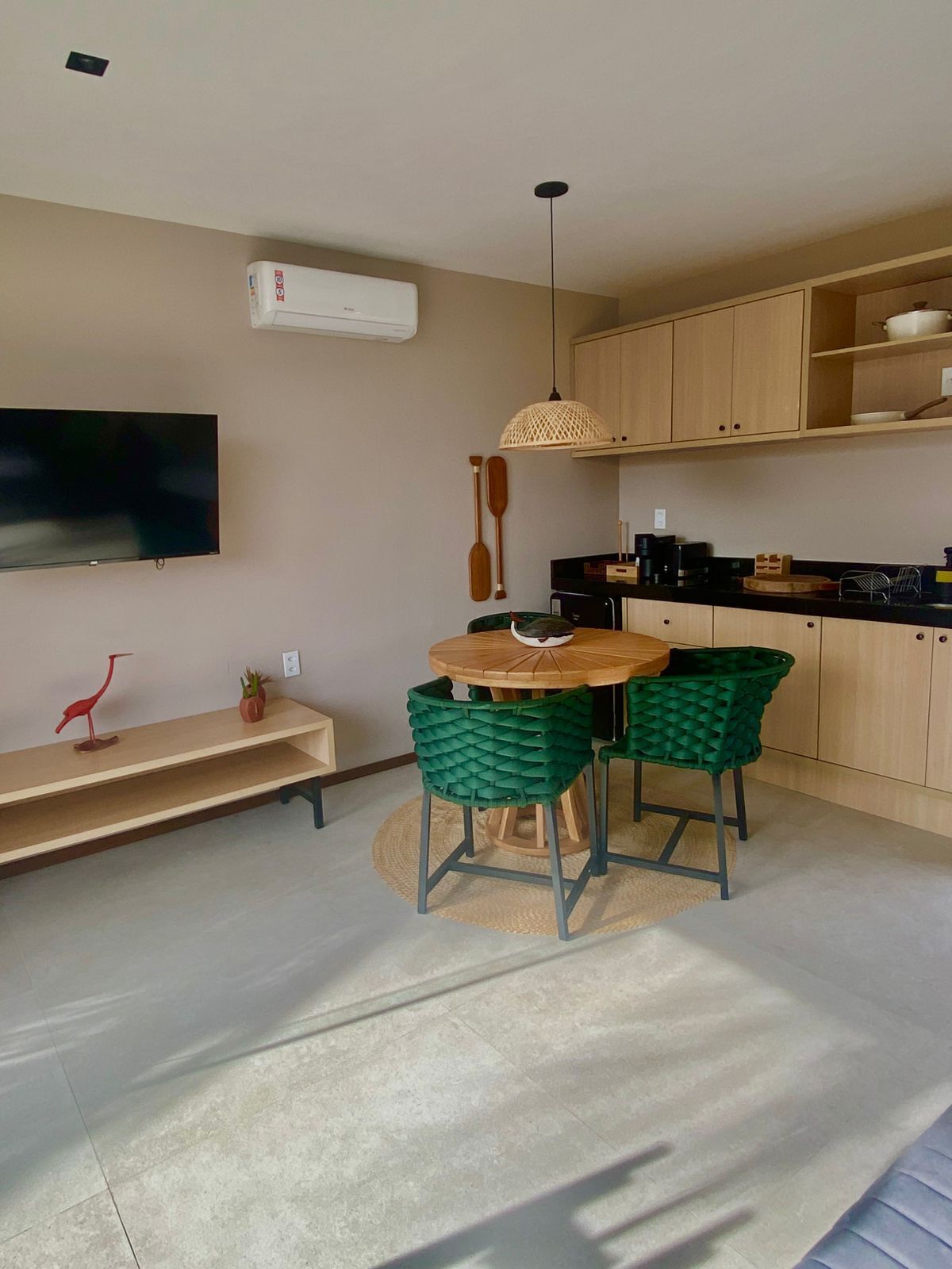A modern kitchen with wooden cabinets, a dining table with two chairs, a TV mounted on the wall, and a small shelf with decorative items.