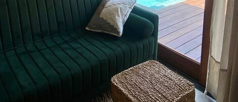 A green sofa with a cushion, a framed map, and a wicker ottoman in a room with a view of a pool and outdoor area.