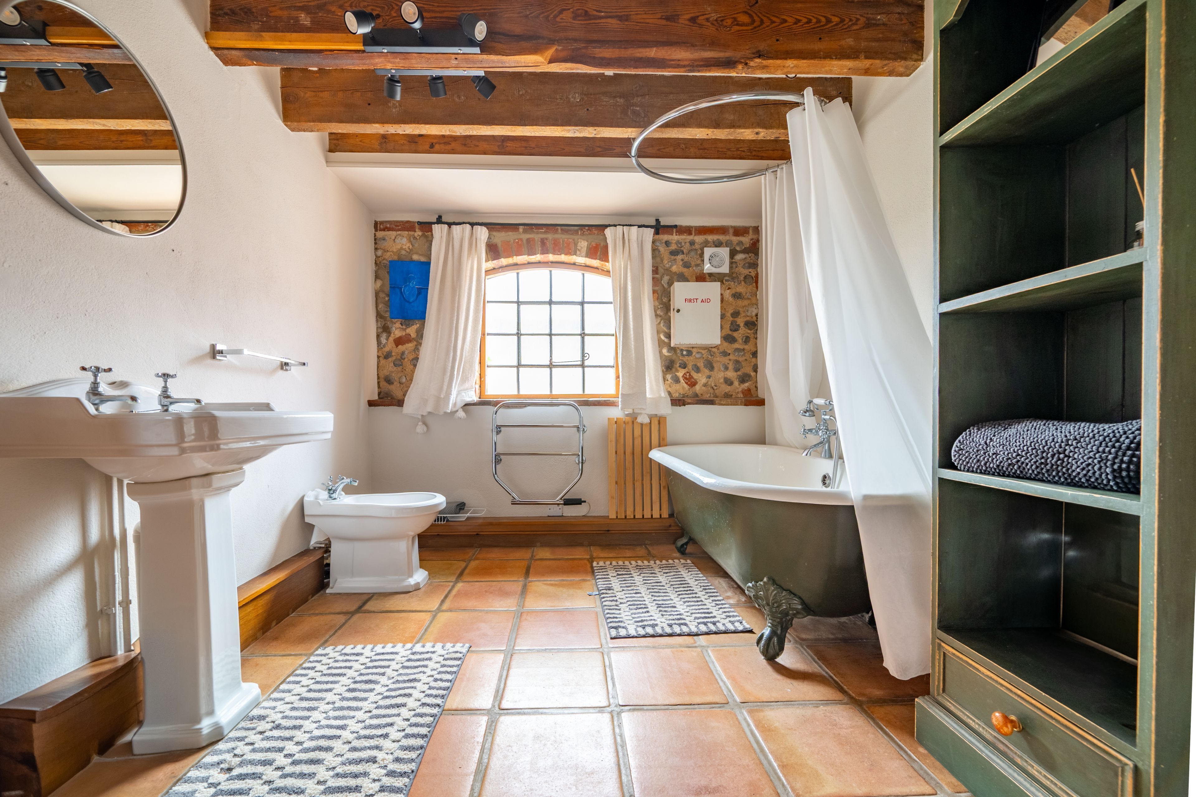 Une salle de bain avec un lavabo sur colonne, des toilettes et une baignoire. On y trouve une fenêtre avec des rideaux, un porte-serviettes et une étagère verte sur laquelle est posée une serviette.