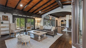 A spacious living room with a high wooden ceiling, large windows, and a cozy seating area with a coffee table and ottomans.