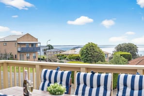 A balcony with striped cushions, a table with a bottle, and a view of the sea and buildings.