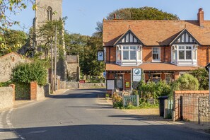 A traditional brick building with a gabled roof, a small shop on the ground floor, and a church tower in the background. - Liz's Lodge, Weybourne, Norfolk (Weybourne)