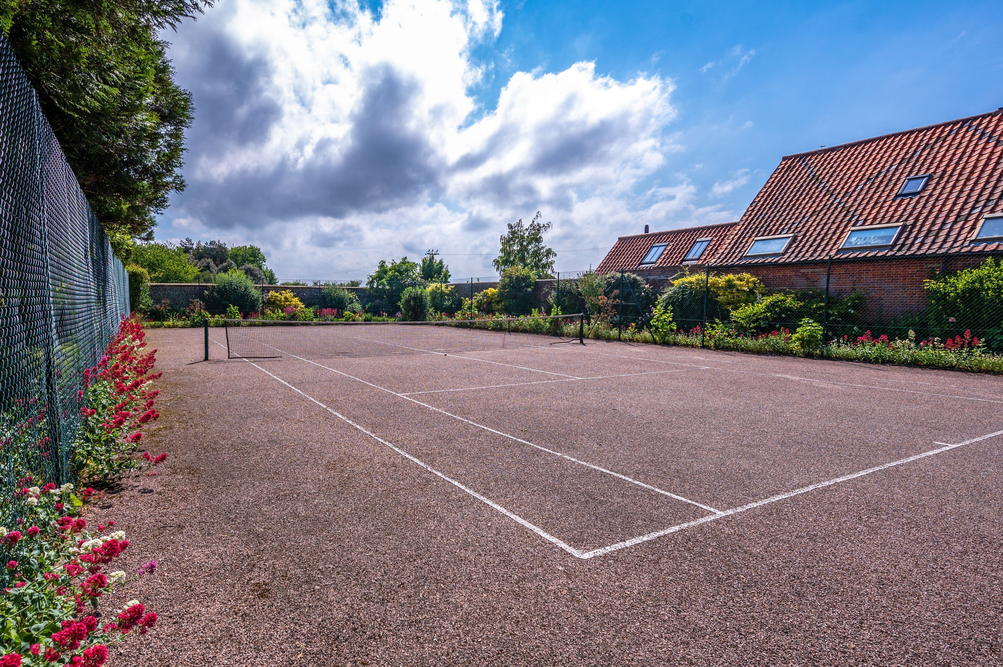 Un court de tennis en plein air, doté d’un filet, entouré d’une clôture et agrémenté de plantes en fleurs.