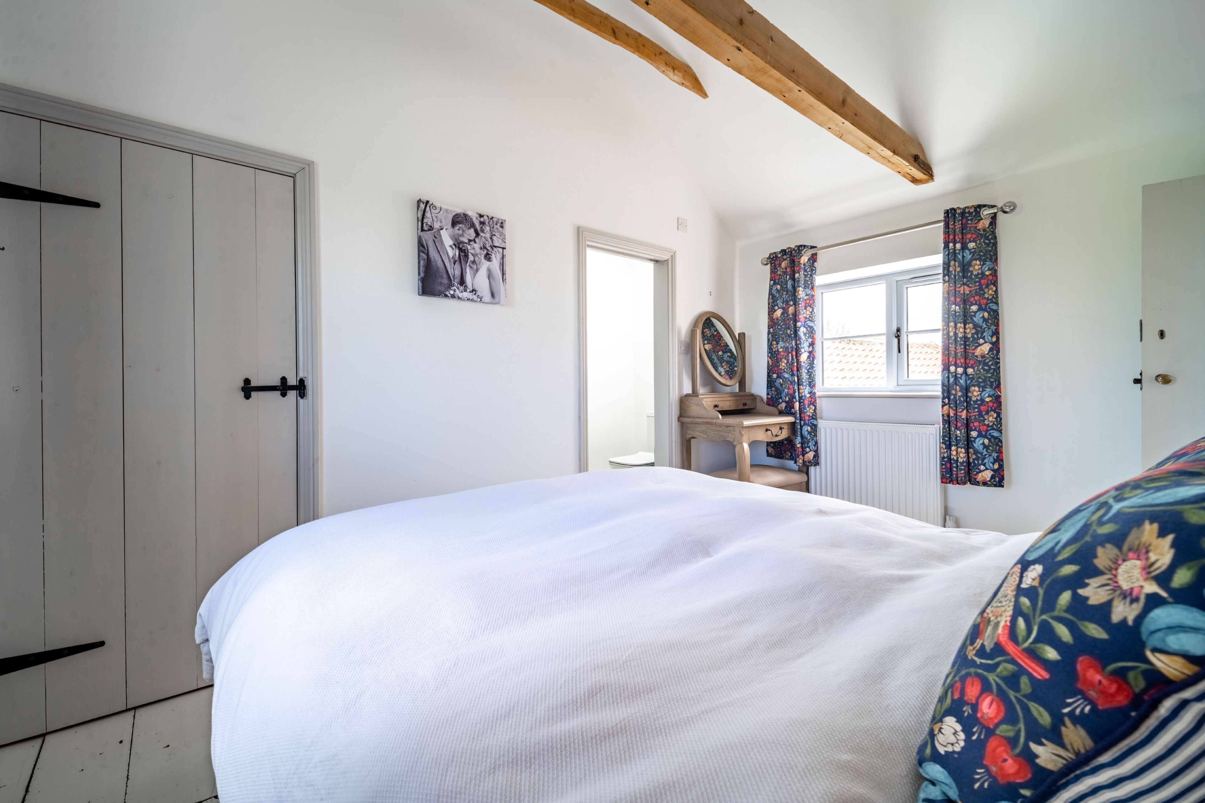 Une chambre à coucher avec un grand lit, une poutre en bois au plafond, une commode, un miroir et une fenêtre ornée de rideaux à motifs floraux.