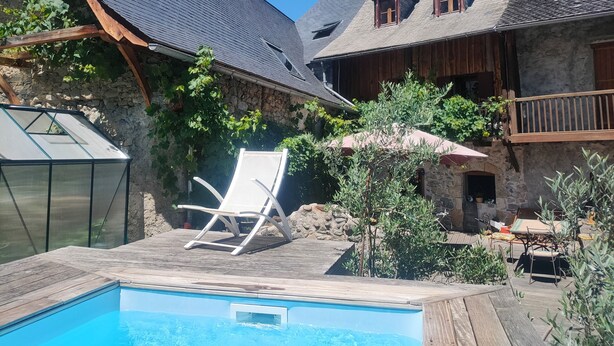 A pool area with a wooden deck, a white lounge chair, and a stone building with a sloped roof. - Les Chatougoulis (Bordes-Uchentein)