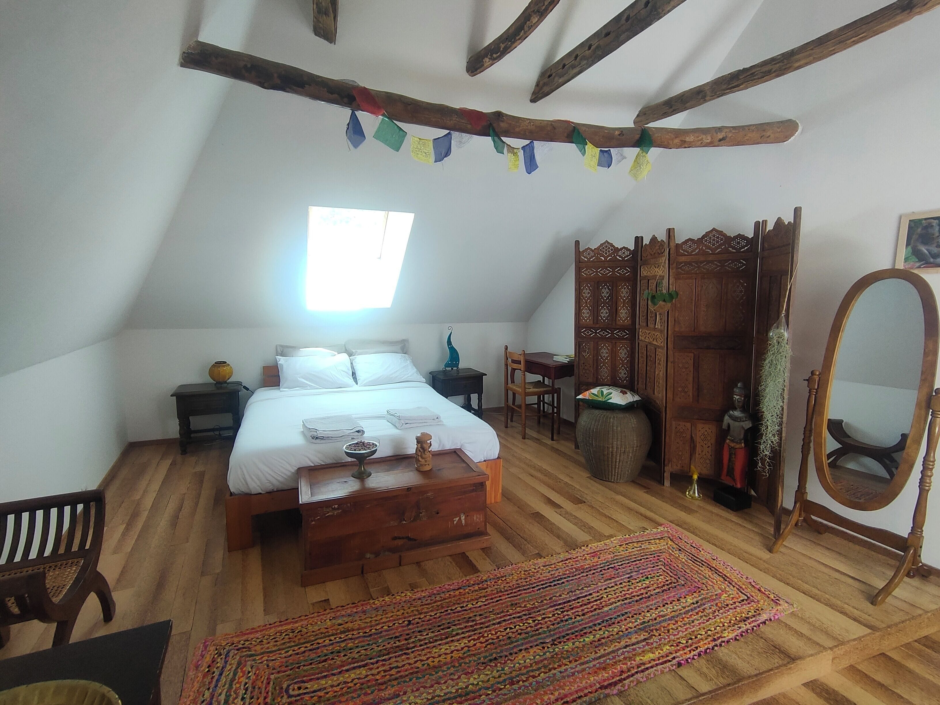 A bedroom with a sloped ceiling, a bed with white linens, a wooden wardrobe, a mirror, and a colorful rug.