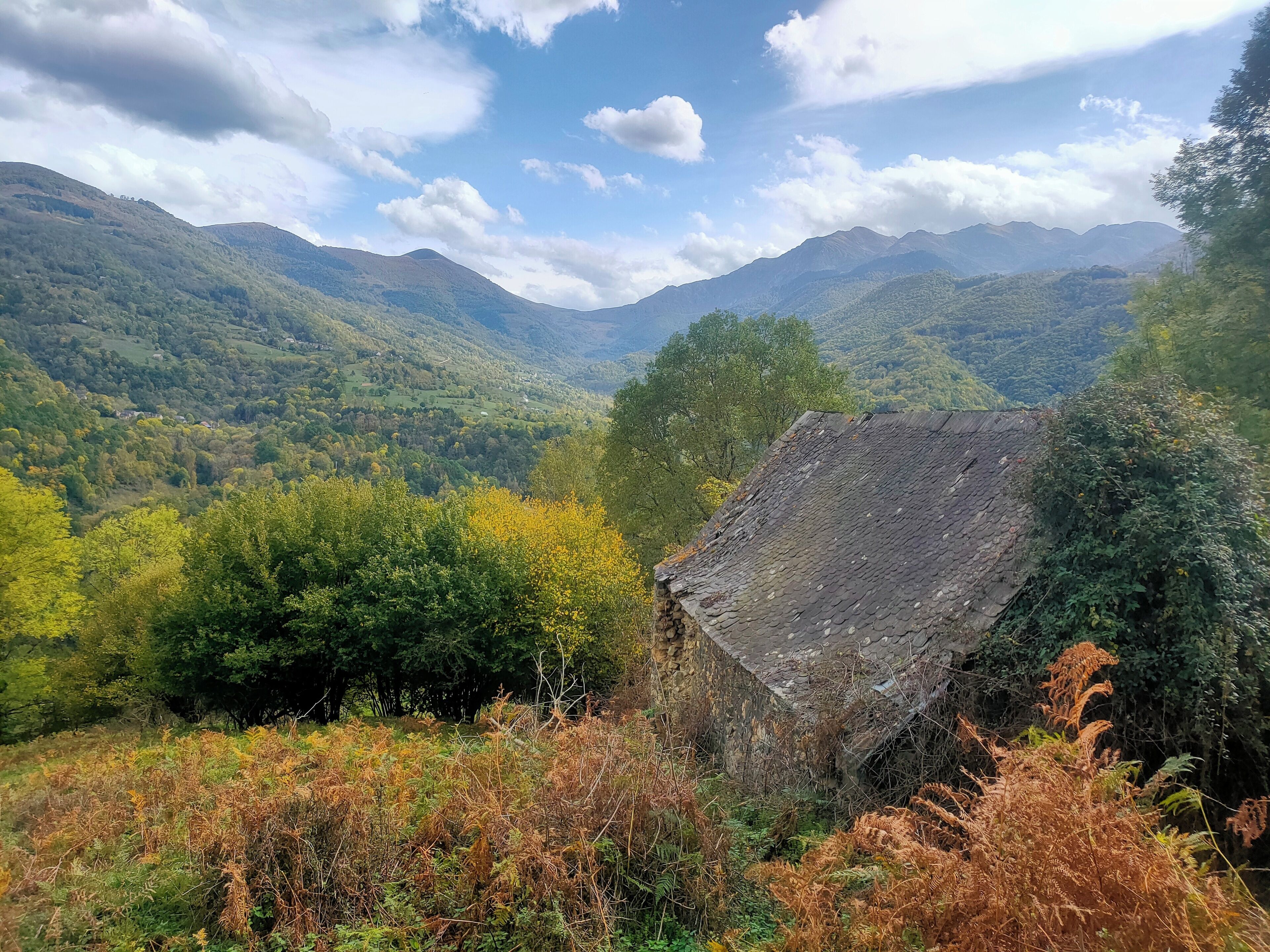 Ländliche Landschaft mit einem Steinhaus, umgeben von üppigem Grün und Bergen im Hintergrund.