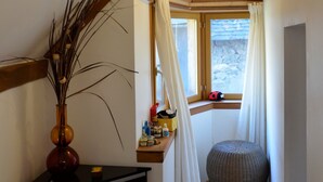 A narrow hallway with a wooden ceiling, a small window with curtains, a vase with branches, a scale, and a round ottoman. - Les Chatougoulis (Bordes-Uchentein)