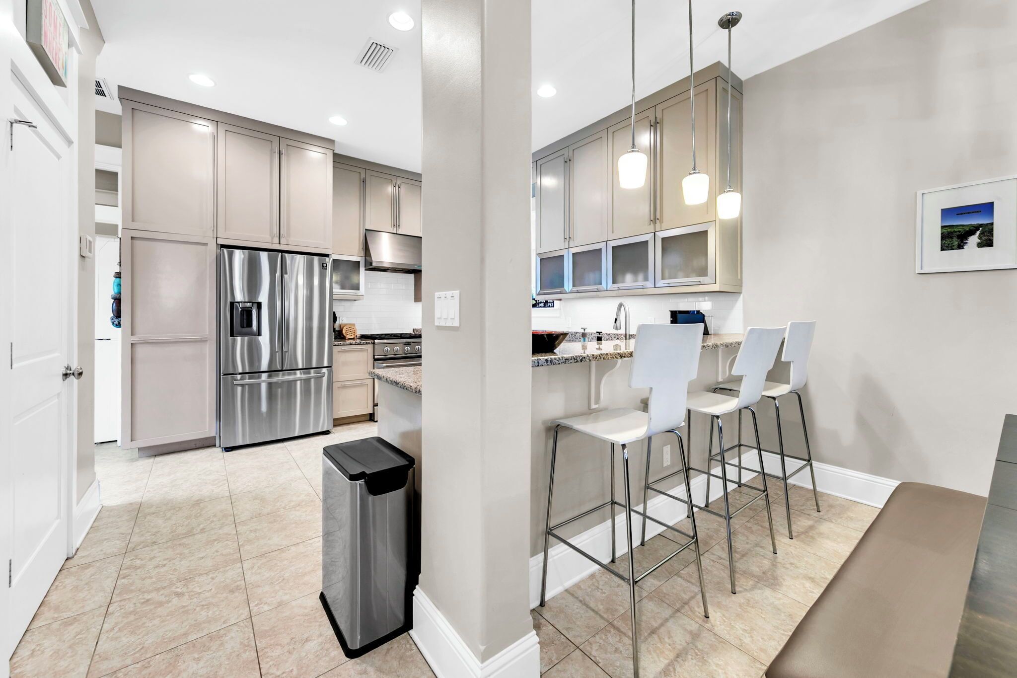 A modern kitchen with stainless steel appliances, a trash bin, and a bar area with white chairs.