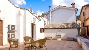 Un patio con sillas de mimbre y una mesa, un edificio blanco con techo de tejas y una unidad de aire acondicionado.