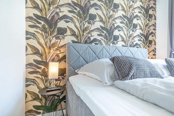 A neatly made bed with a grey headboard, a bedside lamp, and a small round table with a remote control.