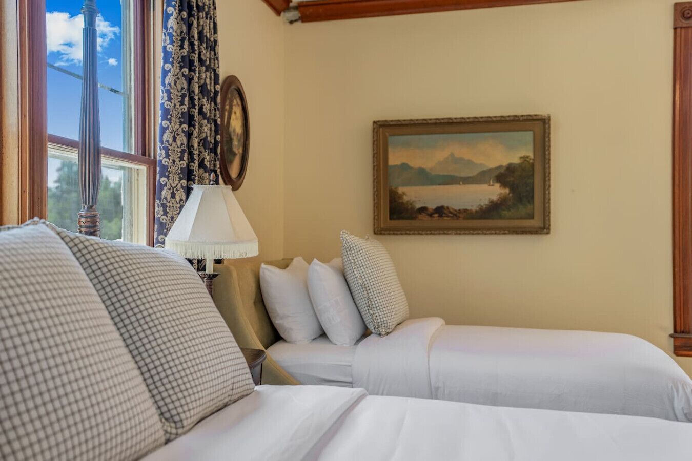 A bedroom with two beds, a lamp, a painting, and a window with curtains.