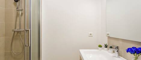 Une salle de bain moderne avec une douche à l’italienne en verre, un lavabo blanc avec un miroir et un vase rempli de fleurs bleues.