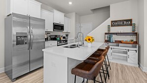 Cocina moderna con armarios blancos, electrodomésticos de acero inoxidable, una isla central con taburetes y un letrero que indica una barra de café.