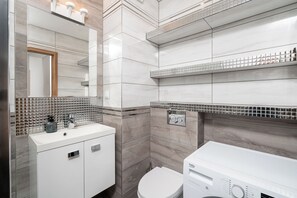 A modern bathroom with a white sink, mirror, and toilet. There are shelves with toiletries and a washing machine. - AS HOME Tęczowa 89 (Wrocław)