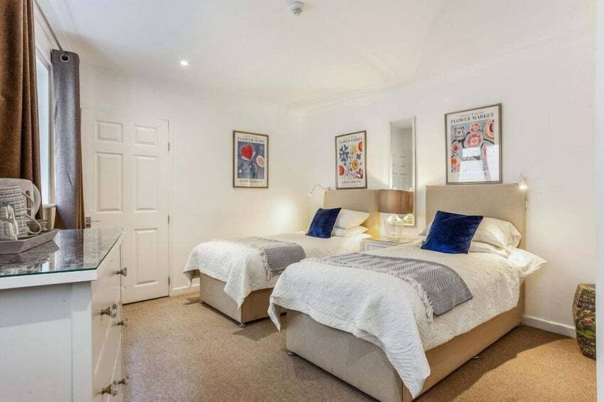 A bedroom with two beds, a desk, and a chair. There are framed pictures on the wall and a window with curtains.