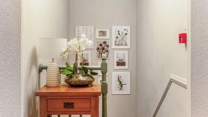 Un corridoio con una consolle in legno che ospita una lampada, un vaso con fiori e una ciotola decorativa. Le pareti sono decorate con quadri incorniciati e un segnale di uscita di sicurezza rosso.