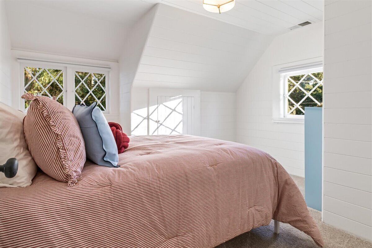 Un dormitorio con techo inclinado, cama con colcha a rayas y dos ventanas con marcos blancos y vidrios en forma de rombo.