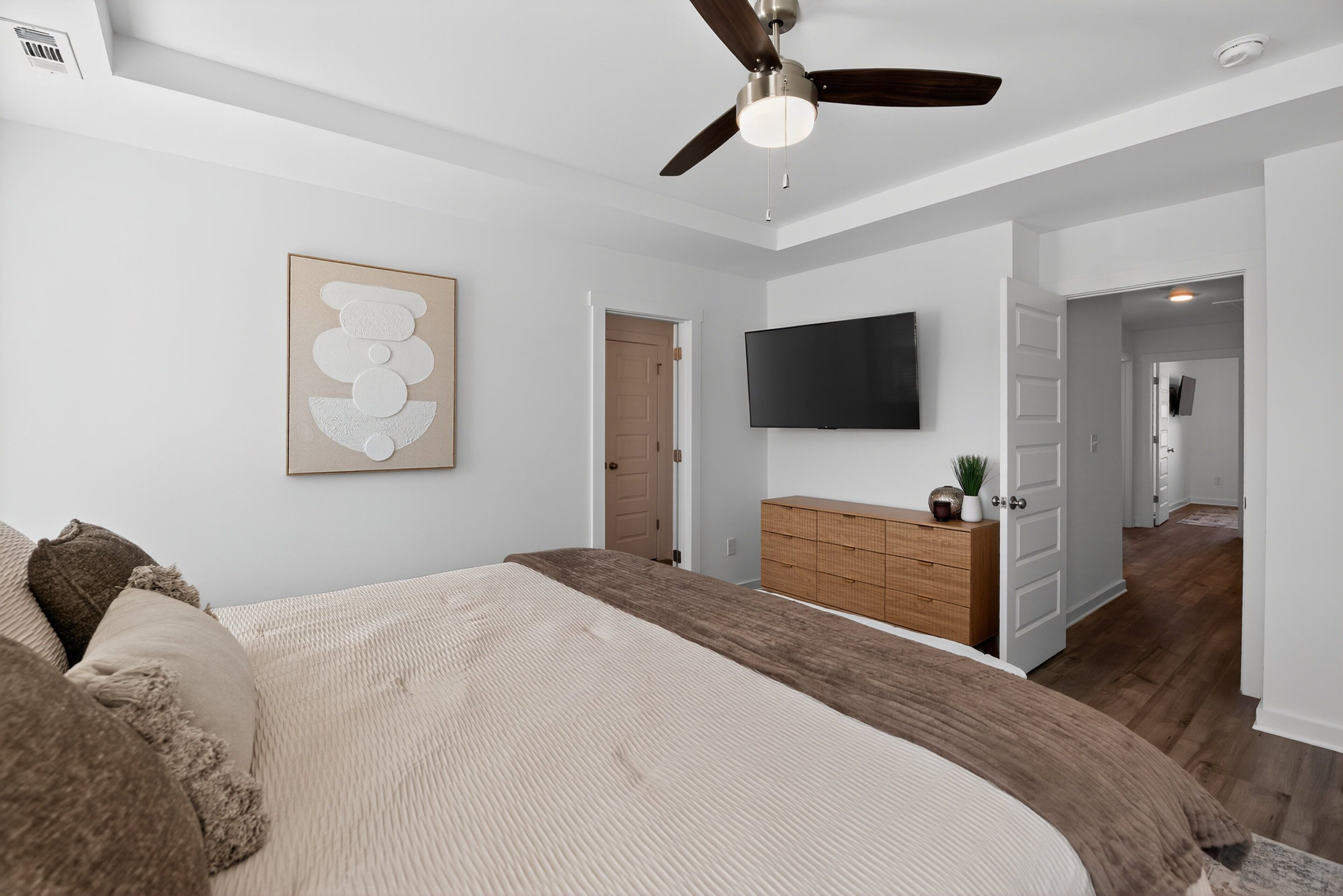 A bedroom with a bed, a ceiling fan, a TV, a wooden dresser, and a door leading to another room.