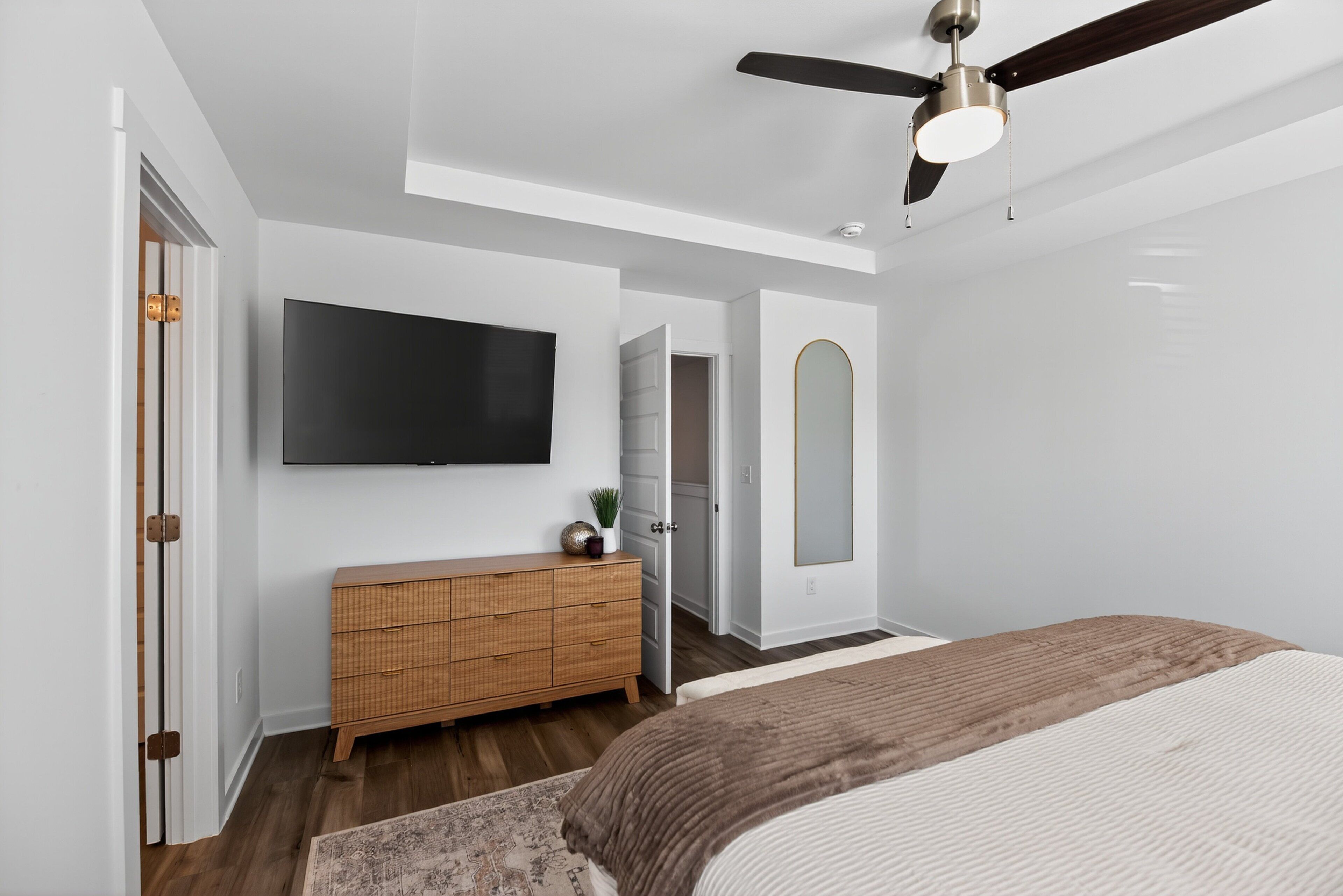 A modern bedroom with a flat-screen TV, a ceiling fan, a wooden dresser, and a large bed.