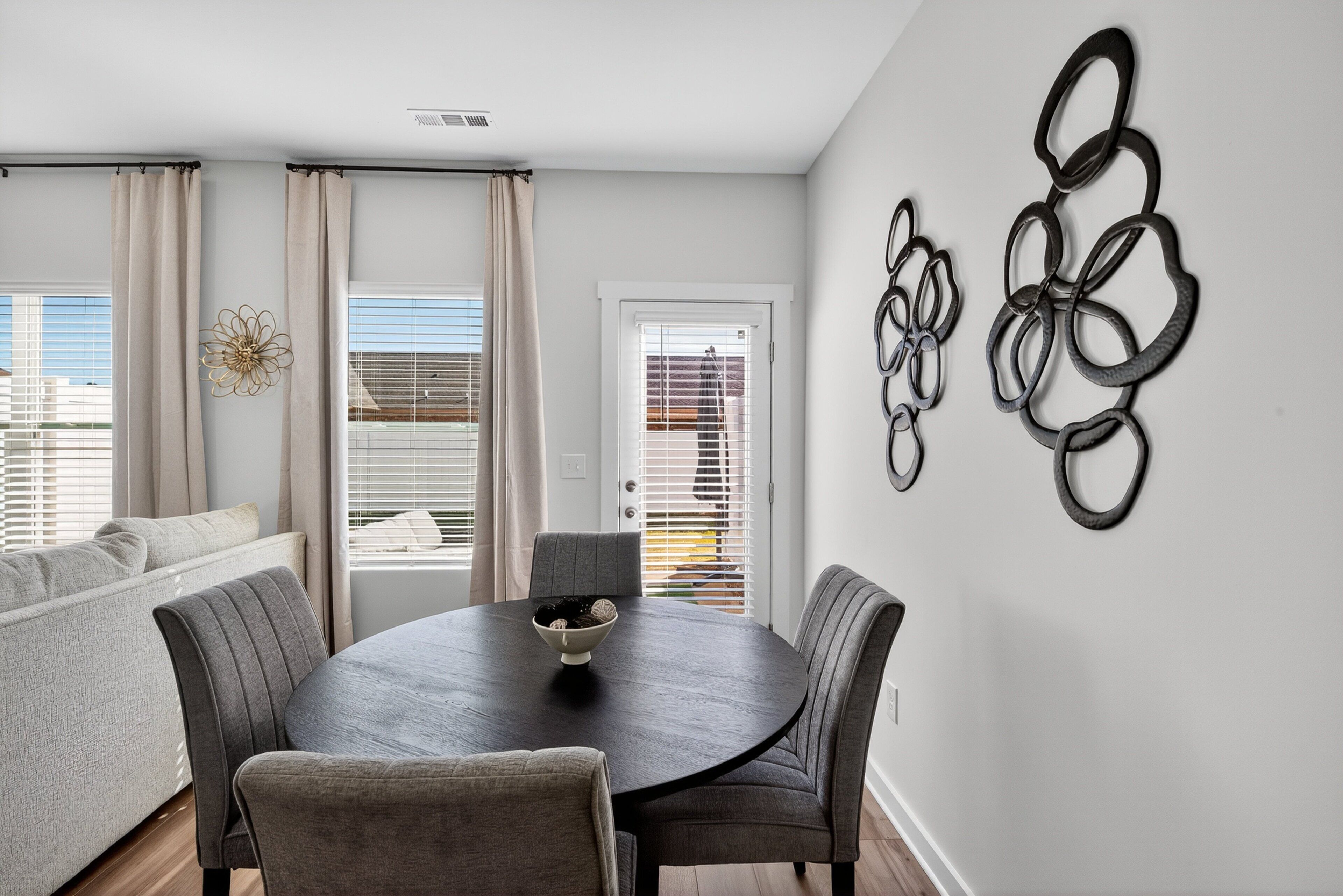 A dining area with a round table, grey chairs, and a bowl of cookies. There are curtains, a door, and a window with blinds.
