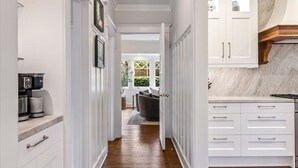 A narrow hallway with white cabinetry, a kitchen area with marble countertops, and a view into a living room with a dark sofa and a window.