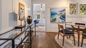A modern dining area with a glass table, wooden chairs, and a painting of a coastal landscape.