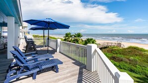 A wooden deck with lounge chairs and a blue umbrella, overlooking a beach with palm trees and a wooden walkway leading to the shore. - Casa Azul 3 Bedroom Home (Port Bolivar)