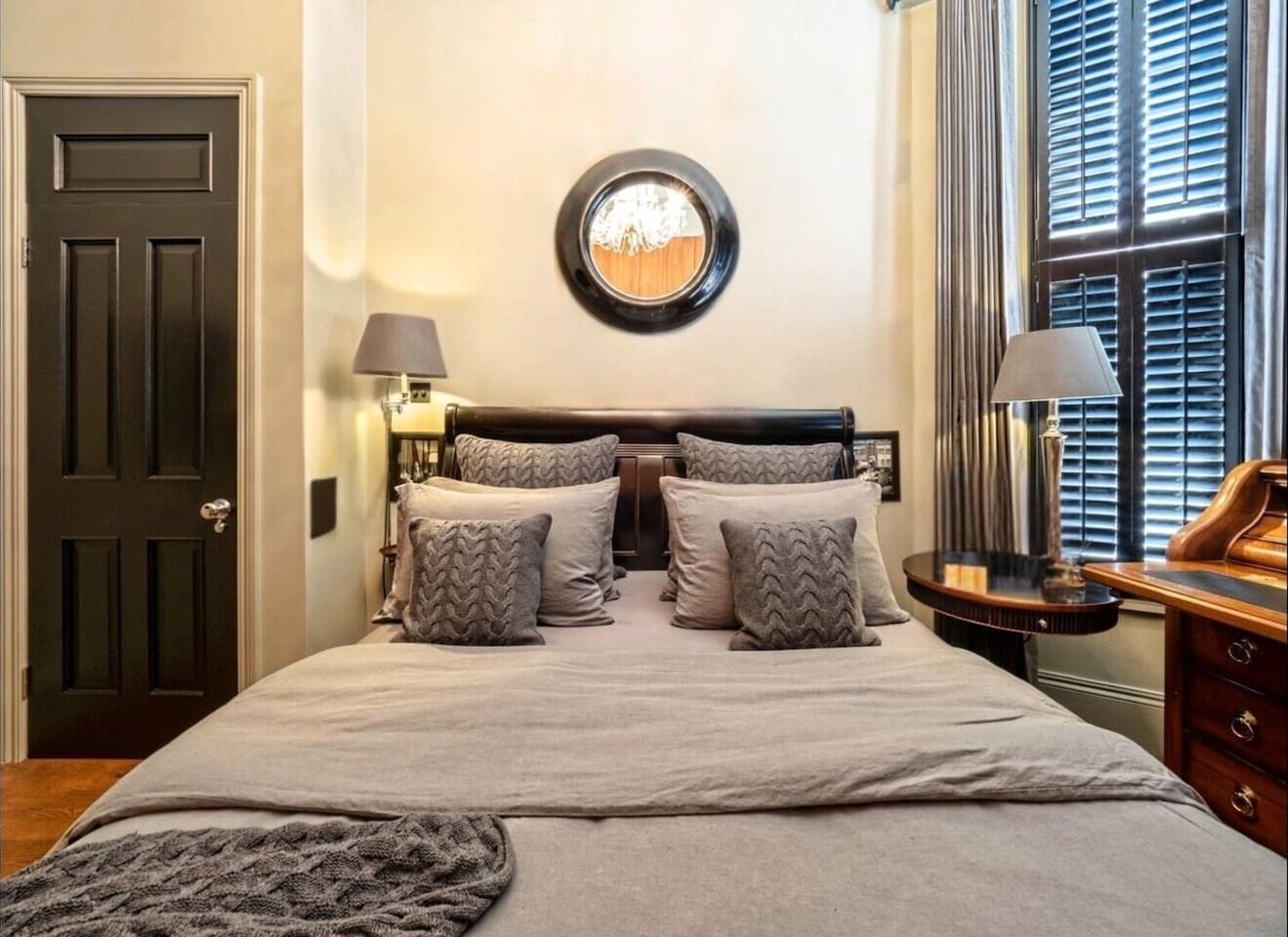 A bedroom with a double bed, two bedside tables, a mirror, and a window with blinds.