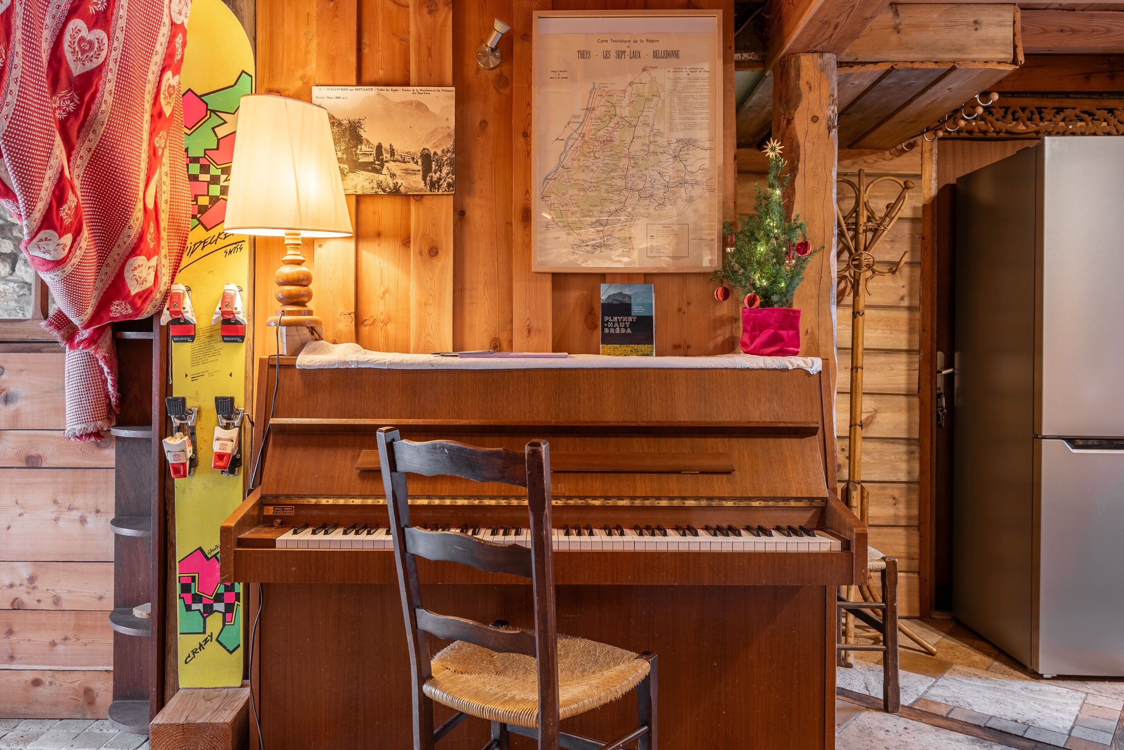 A wooden interior with a piano, a chair, a map on the wall, and a small Christmas tree.