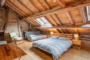 Un accogliente sottotetto adibito a camera da letto, con soffitto spiovente, travi in legno, un letto con lenzuola a righe, una sedia e un tavolino con una lampada.