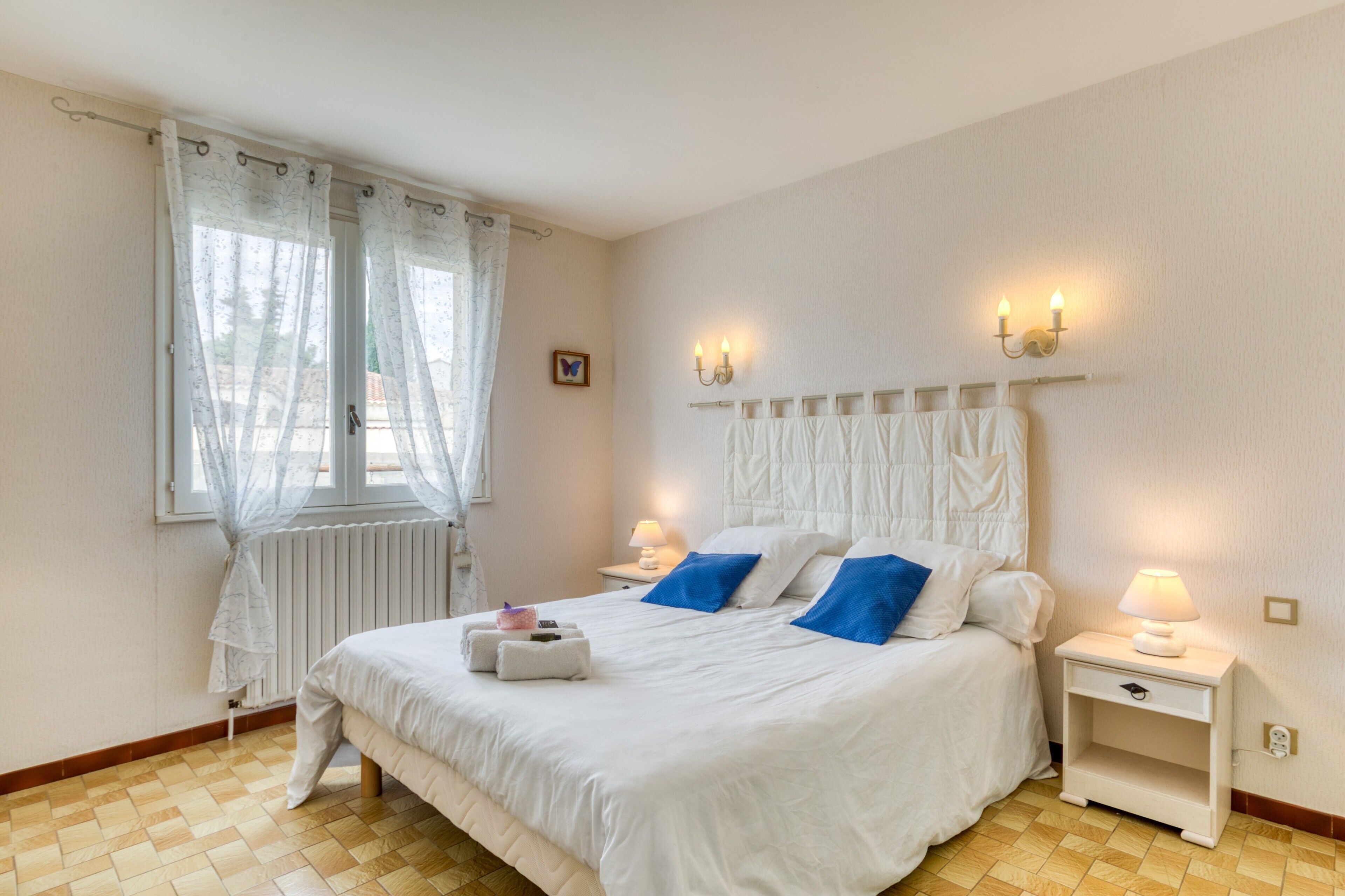 Ein Schlafzimmer mit einem Bett, zwei Nachttischen mit Lampen, einem Fenster mit Vorhängen, einem Heizkörper und einer Wandleuchte.