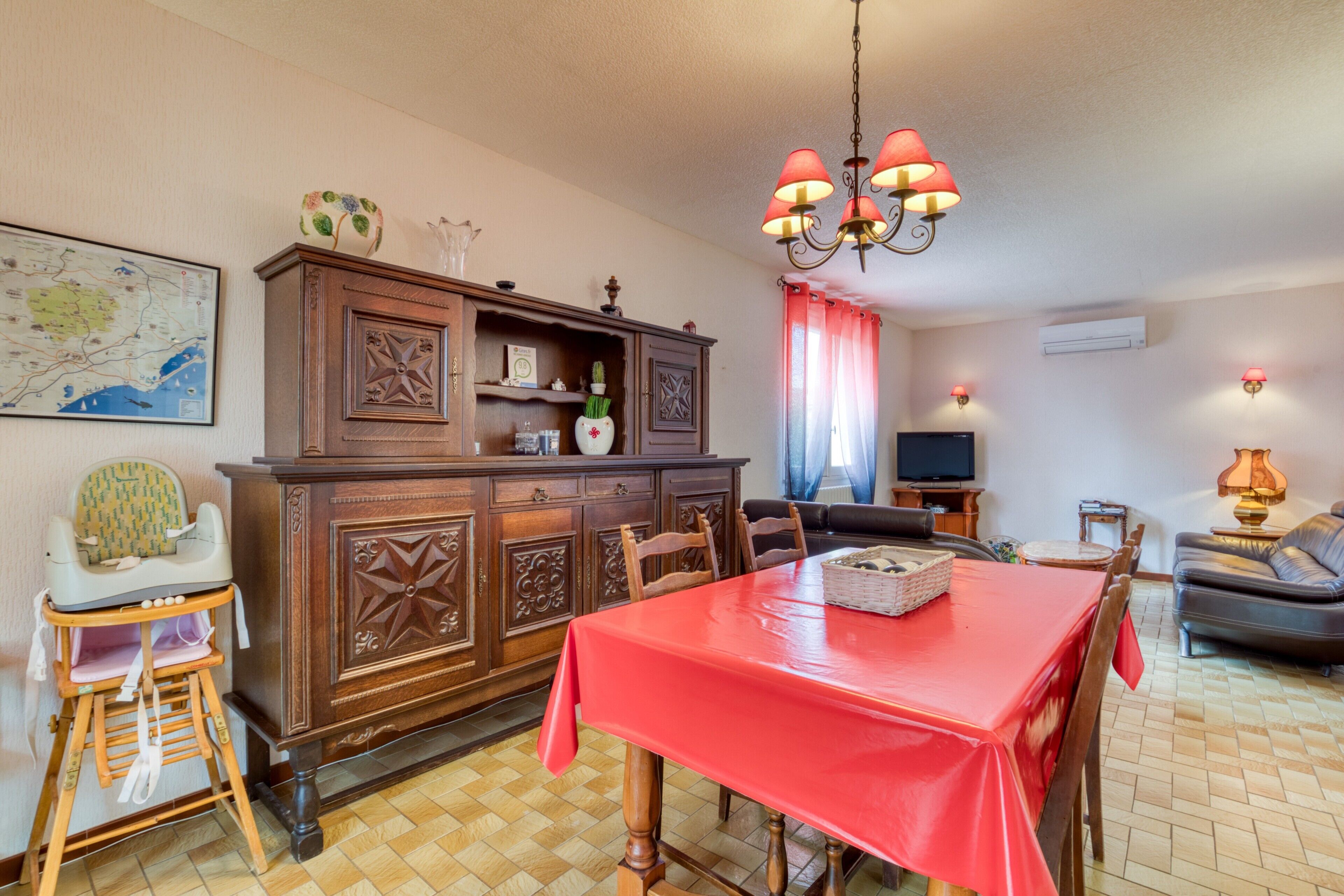 A dining area with a red tablecloth, wooden chairs, a high-backed dining table, a wooden cabinet with decorative carvings, a chandelier, a television, a sofa, and a map on the wall.
