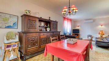 A dining area with a red tablecloth, wooden chairs, a high-backed dining table, a wooden cabinet with decorative carvings, a chandelier, a television, a sofa, and a map on the wall.