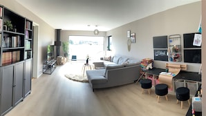 Un salon moderne avec un canapé gris, une bibliothèque, un bureau avec une chaise et une grande fenêtre donnant sur l’extérieur.
