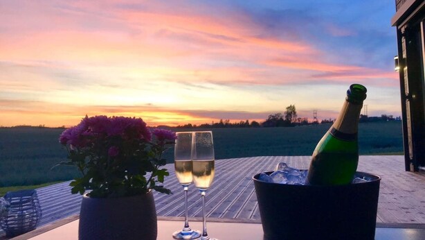 Une table avec une bouteille de champagne, deux verres et une plante en pot, le tout placé sur une terrasse en bois donnant sur un champ au coucher du soleil.