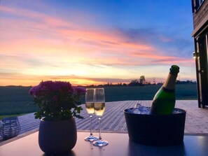 A table with a bottle of champagne, two glasses, and a potted plant on a wooden deck overlooking a field during sunset. - Modern detached house with sauna! 6 beds. 20 min. From Oslo. (Kråkstad)